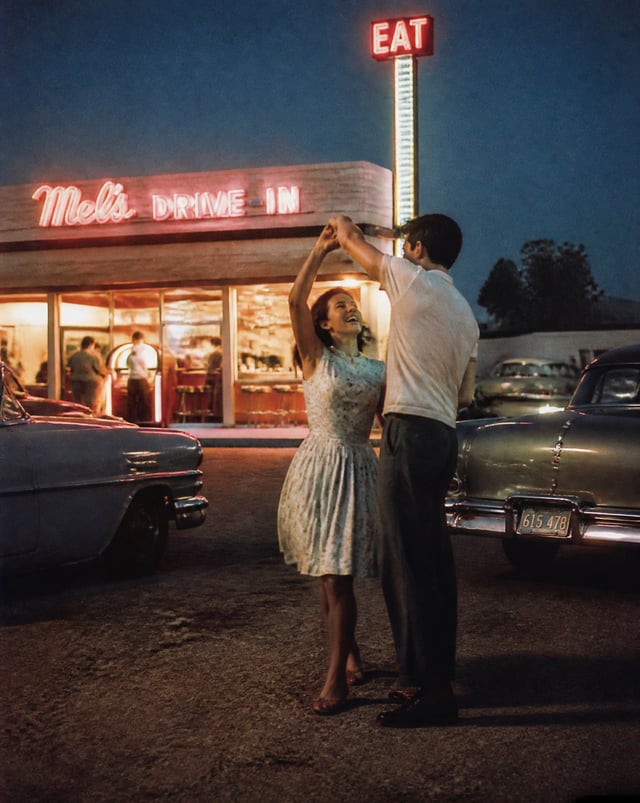 1956 roadside diner at blue hour, a young couple spontaneously slow dancing beside a glowing jukebox