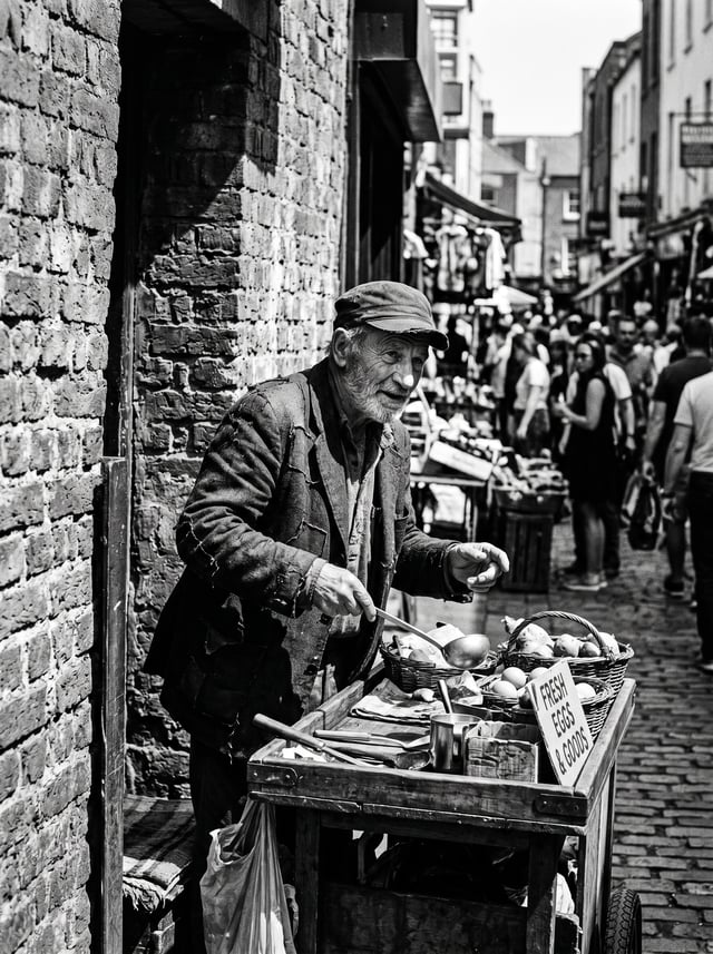 Candid black and white street photography portrait of [an elderly street vendor] framed by a brick w
