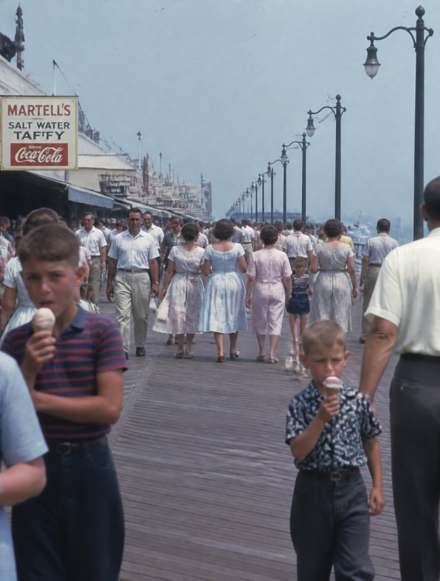 1950s seaside boardwalk filled with families, children eating ice cream, couples strolling, candid m