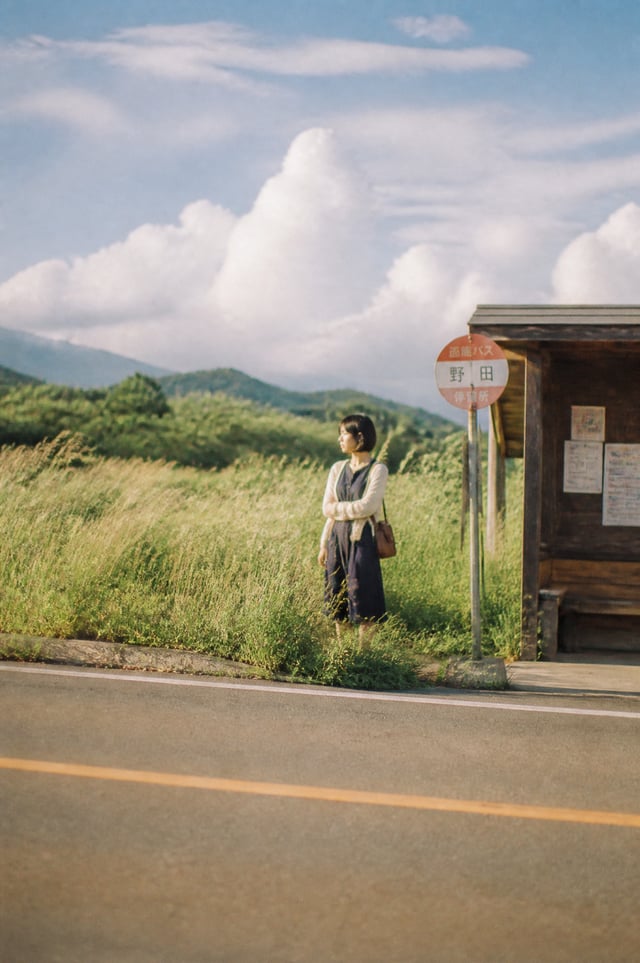 A beautiful Japanese woman in her early twenties waiting alone at a quiet countryside bus stop durin