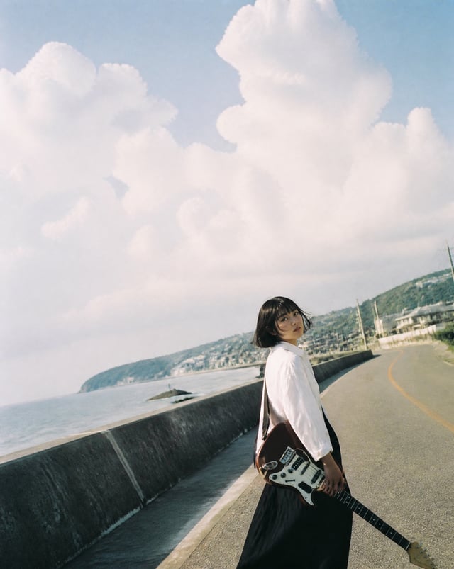 A beautiful Japanese woman in her early twenties standing alone on a quiet seaside road carrying a v