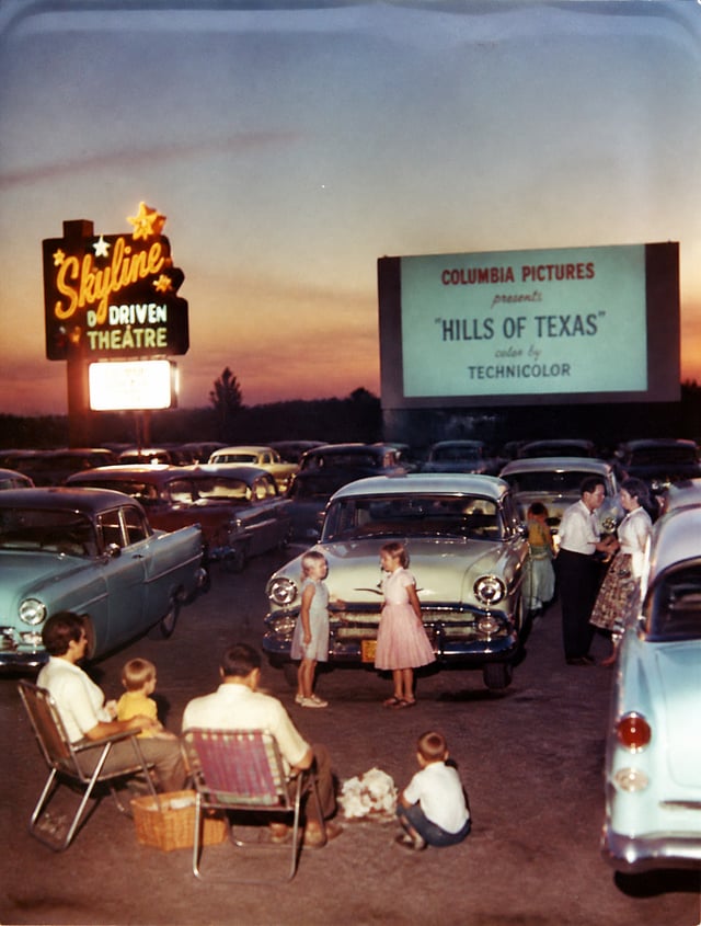 1950s drive-in movie theater at dusk, rows of classic cars, families sitting outside their vehicles,