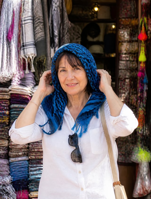 A middle-aged woman with a gentle smile poses in front of a densely packed market stall, her hands r