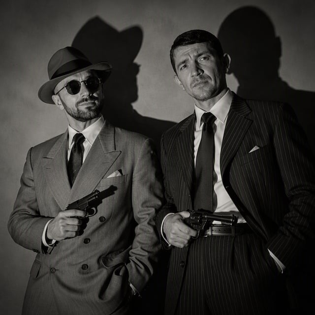 Two sharply dressed men stand tensely in a stark mid-century noir interior, the left figure in a lig