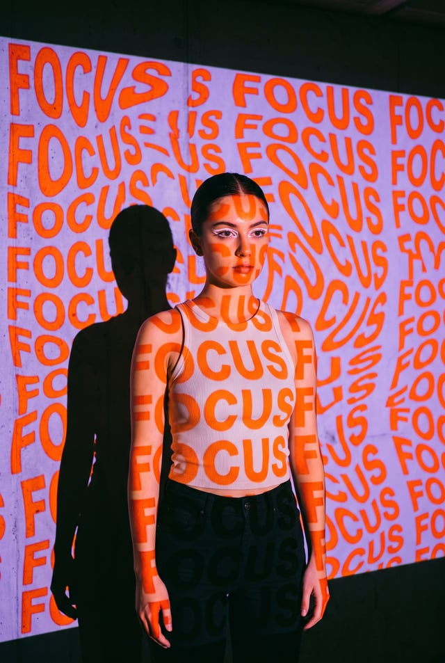 A young woman stands in front of a wall with a projection of the word "FOCUS" repeated in a distorte