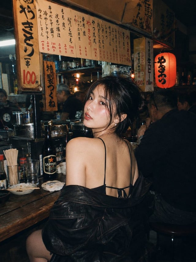 A beautiful young East Asian woman with long dark wavy hair and striking makeup, sitting at a wooden