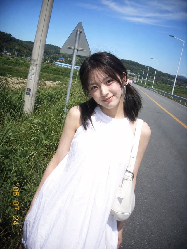 复古 CCD 卡片机夏日旅行抓拍，一位20岁出头的年轻东亚女idol站在乡间公路边。黑棕色低位双马尾，细碎刘海，浅粉色发圈，淡妆，冷白皮，真实皮肤。白色无袖高领宽松连衣裙，轻薄棉质，右肩白色单肩包。身