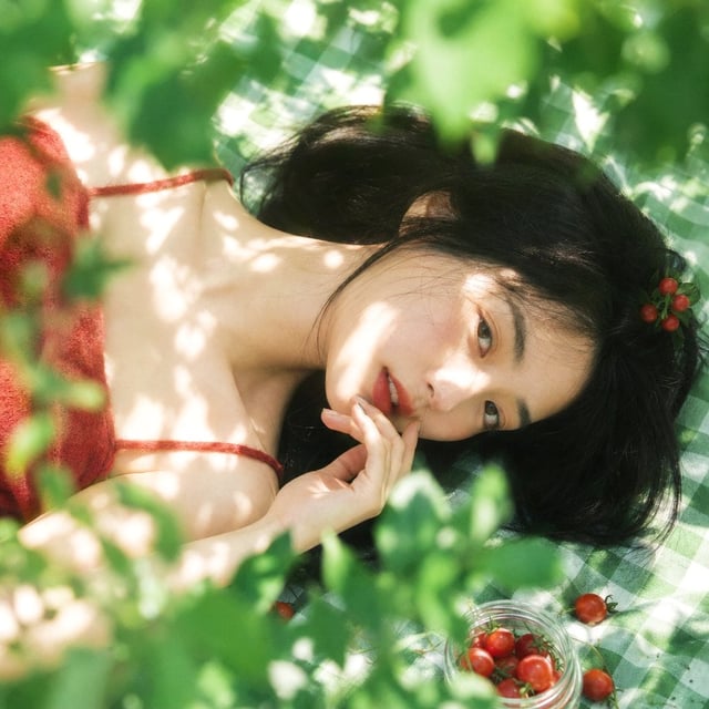 真人摄影，夏日植物系生活方式写真，清透日系胶片感，竖幅 2:3，俯拍近景到面部特写，年轻女性横向躺在绿白格纹野餐布上，脸部位于画面中部偏右下作为主视觉，周围绿植前景占较大比例并形成柔和遮挡，底部少量透