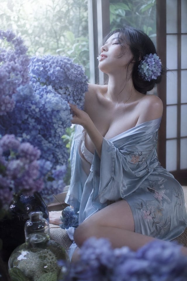 真人摄影，日系柔焦胶片感花卉写真，竖幅 2:3，窗边自然逆光与轻微过曝光晕。日系美女，身材夸张，偏右侧坐，冷白到象牙白肤调受冷蓝窗光轻微偏色，黑色低盘发配蓝紫绣球花发饰，发丝散落；穿浅蓝灰丝缎和服式外