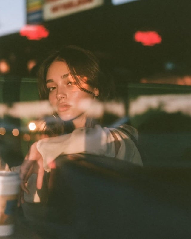 Ultra-cinematic lifestyle portrait photographed through a café window at blue hour, beautiful young 