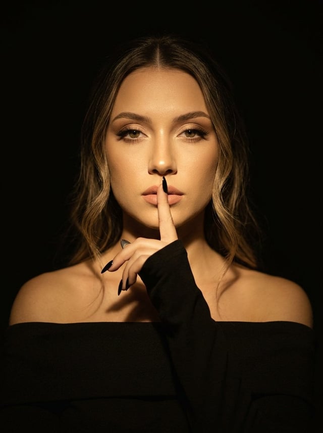 A woman stands in a dark room, her body facing the camera, her shoulders open, one hand raised to he