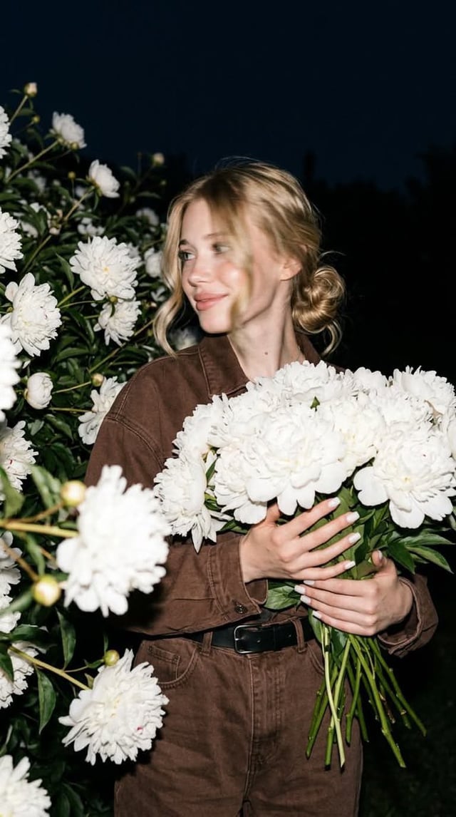 A candid nighttime editorial portrait of a young blonde woman standing in a lush garden filled with 