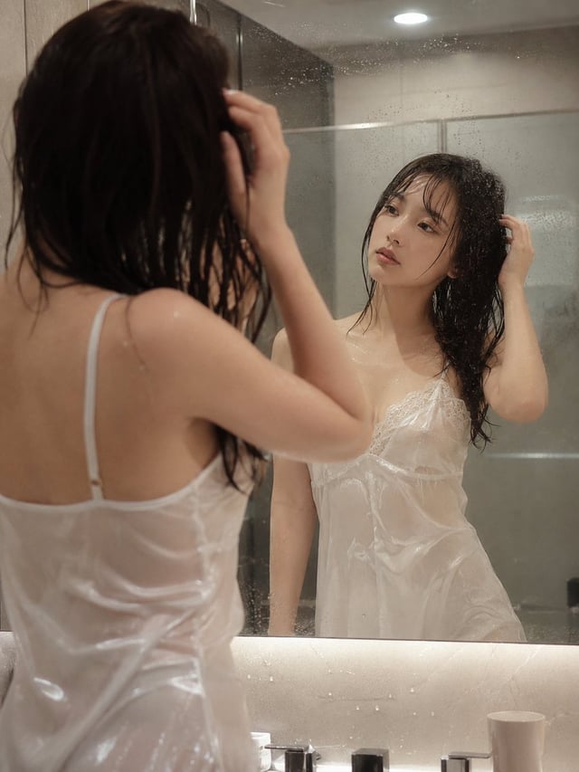 一名23歲日系女性，穿著微濕的白色細肩帶絲綢睡衣。她站在充滿水氣的浴室鏡子前，正隨意整理著微濕的頭髮。水氣讓輕薄的絲綢布料微微貼合在優美的身體曲線上，在浴室柔和的頂光下散發著細膩的光澤與微透感，慵懶且