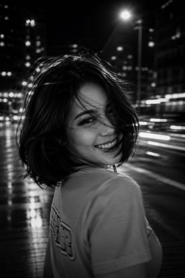 A black-and-white cinematic photograph of a young woman at night in the city, shot with a long expos