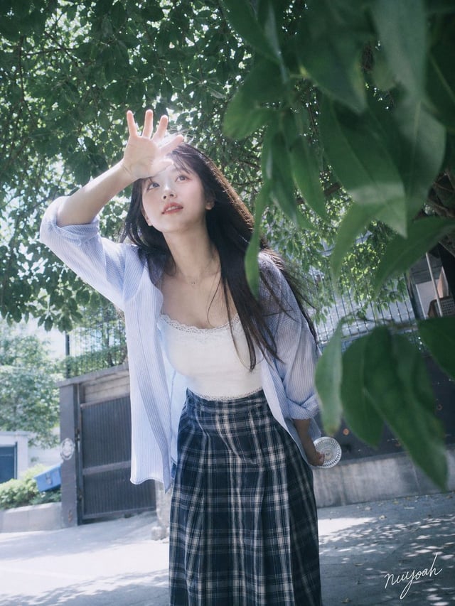 成年年轻女性，清透桃粉甜美妆，深棕长直发与自然风吹碎发，白色蕾丝吊带搭配宽松浅蓝条纹衬衫、深蓝格纹下装；盛夏城市街巷实景，人物处于强烈日照中，皮肤与浅色服装局部高曝光，背景保留冷青色阴影与真实街景杂乱