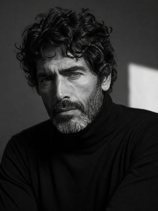 A cinematic black-and-white close-up portrait of a rugged, mature man with thick, naturally curly da